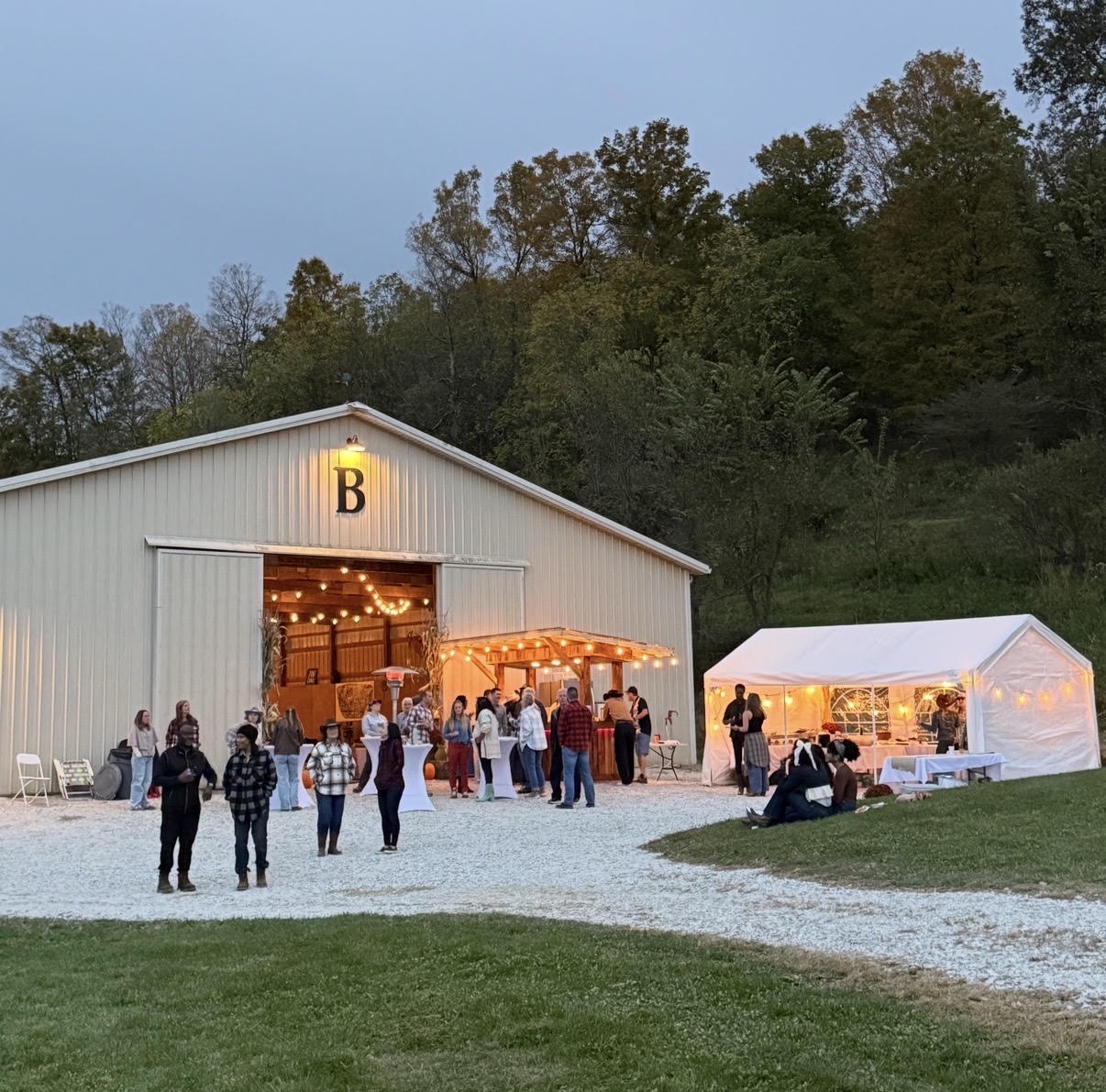 Guests gathering beside a barn and a warmly lit white party tent at dusk