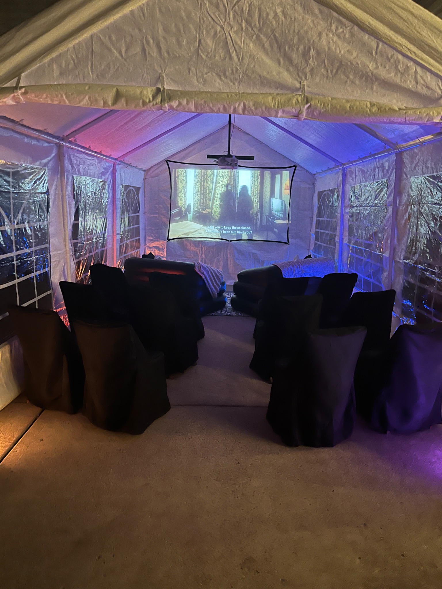 Movie-night tent with a ceiling-mounted projector, screen, inflatable sofas and black-covered chairs under colorful lighting