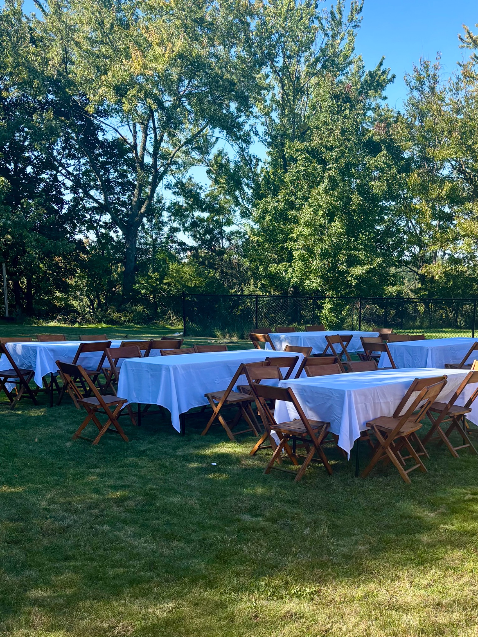 White-covered tables and wooden folding chairs arranged outdoors beneath mature trees