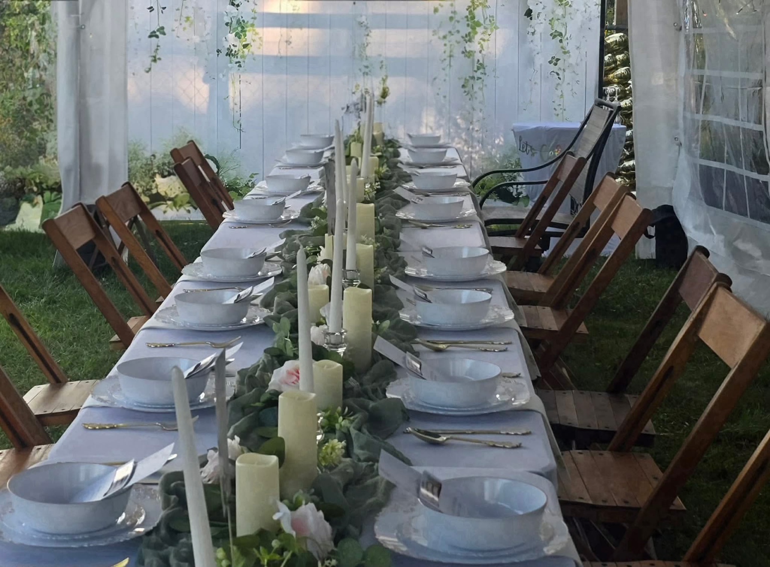 Long outdoor birthday table set with white linens, wooden chairs, greenery, flowers and candles beneath a white tent