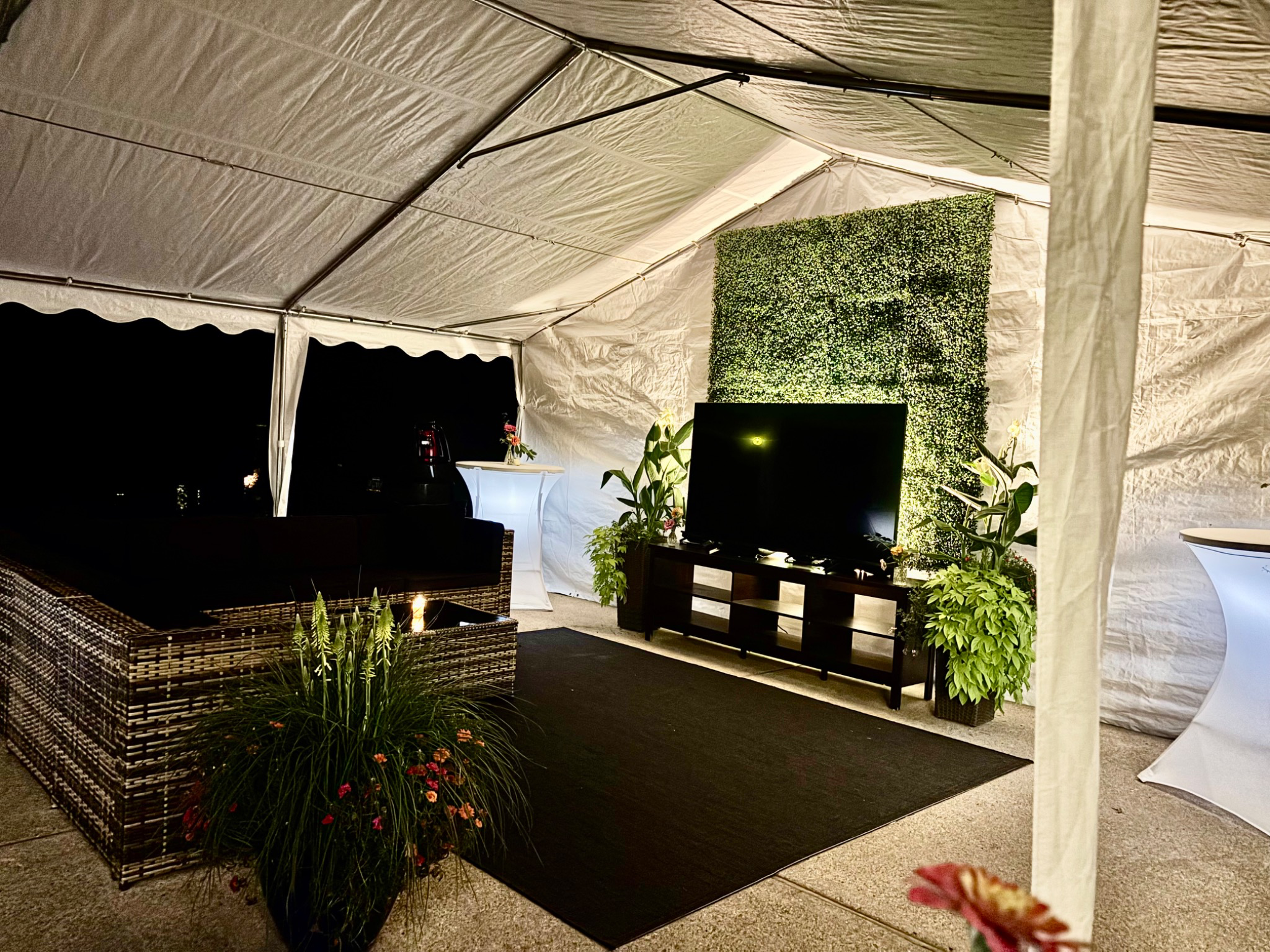 Evening tent setup with softly glowing cocktail tables and an illuminated greenery backdrop