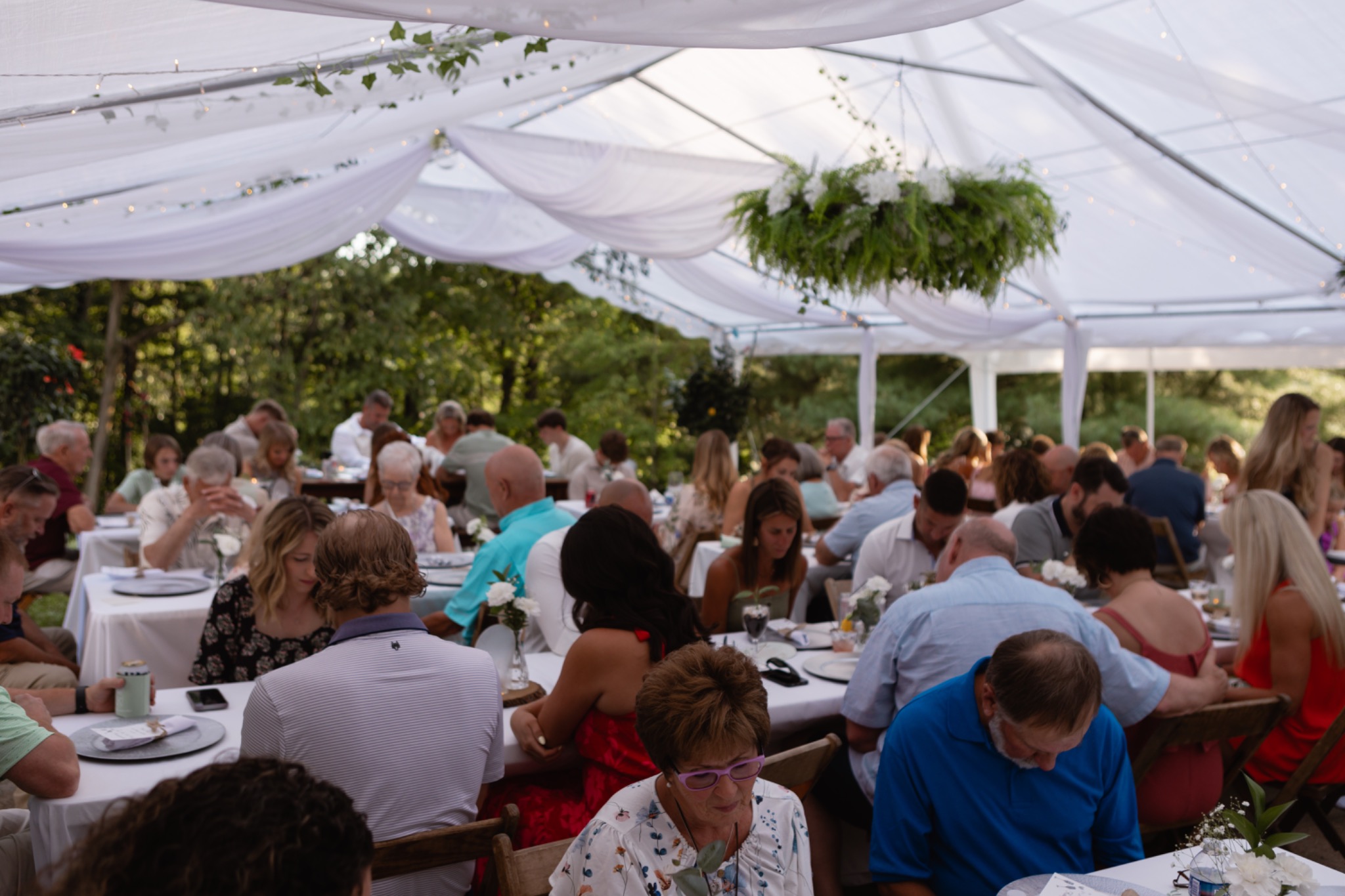 Wedding guests dining at white-covered tables beneath a tent decorated with white drapes, string lights and hanging greenery